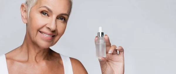 Happy senior woman holding bottle with serum isolated on grey, banner — Stock Photo