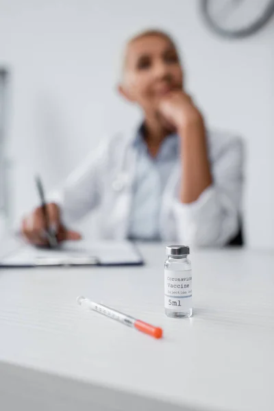 Flasche mit Coronavirus-Impfstoff in der Nähe von Spritze und verschwommenem Arzt im Hintergrund — Stockfoto