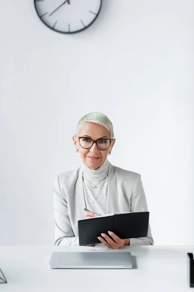 Cheerful senior businesswoman in glasses writing in notebook near laptop — Stock Photo
