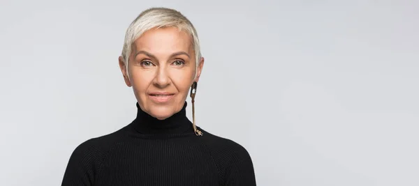 Smiling senior woman with long earring and black turtleneck looking at camera isolated on grey, banner — Stock Photo