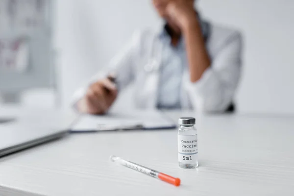 Bottle with coronavirus vaccine and syringe on desk near blurred doctor on background — Stock Photo