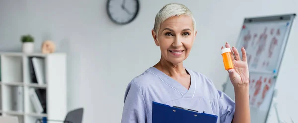 Médecin aîné joyeux tenant presse-papiers et bouteille avec des pilules à la clinique, bannière — Photo de stock