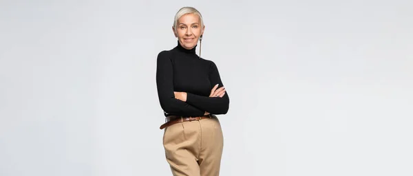 Pleased and trendy senior woman in beige pants and turtleneck posing with crossed arms isolated on grey, banner — Stock Photo