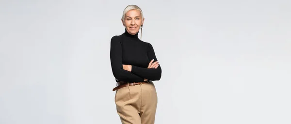 Femme sénior heureuse et tendance en pantalon beige et col roulé posant avec les bras croisés isolés sur gris, bannière — Photo de stock