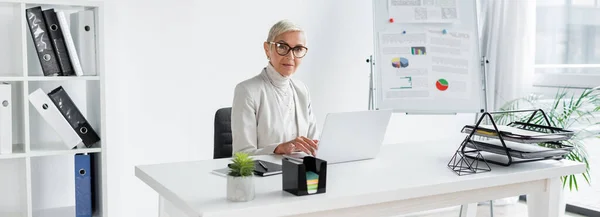 Empresária sênior em óculos sentado na mesa com dispositivos no escritório, banner — Fotografia de Stock