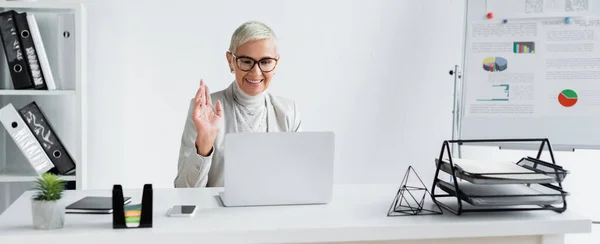 Empresária sênior feliz em óculos acenando a mão durante a chamada de vídeo no laptop, banner — Fotografia de Stock