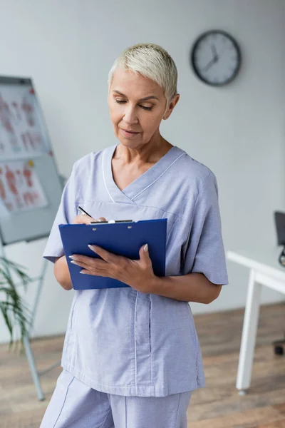 Arzt mit grauen Haaren steht und schreibt Rezept in Klemmbrett — Stockfoto