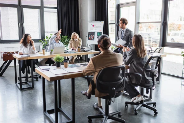 Businessman raising hand during discussion with multiethnic coworkers in office — Foto stock