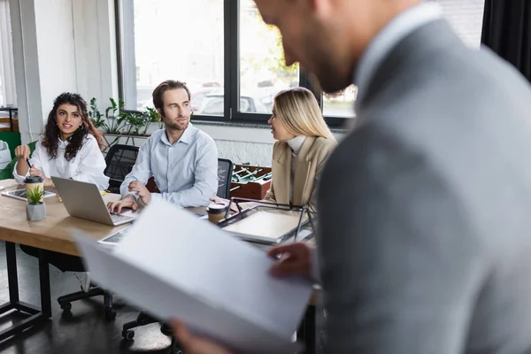 Young business people sitting at desk near man with documents on blurred foreground — Foto stock