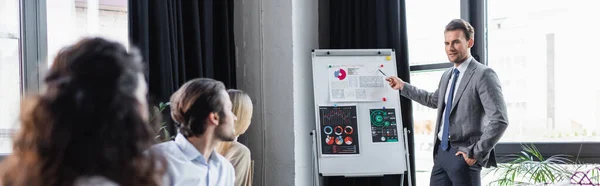 Businessman pointing at flip chart with graphs near colleagues on blurred foreground, banner — Stock Photo