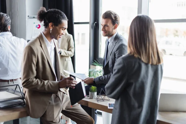 Smiling african american businessman holding book while standing near business partners in office — Stock Photo