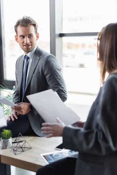 Young businessman in formal wear looking at businesswoman holding documents on blurred foreground — Fotografia de Stock