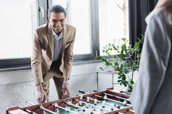 Cheerful african american man in formal wear playing table football with blurred business colleague in office — Foto stock
