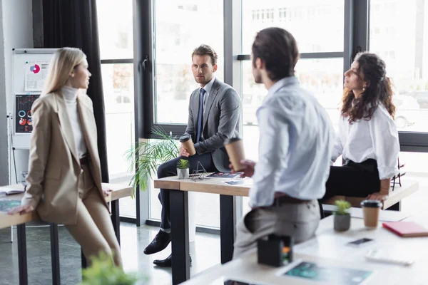 Young business people in formal wear talking while sitting on desks during coffee break — Stock Photo