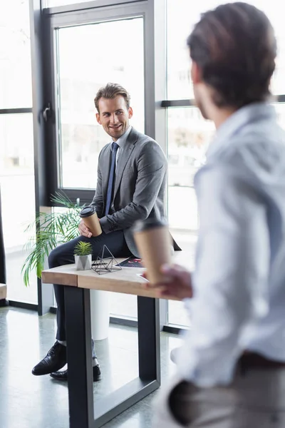 Smiling businessman with coffee to go sitting on desk near blurred colleague — Stock Photo