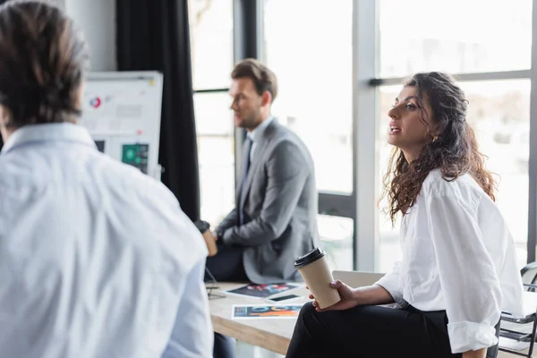 Businesswoman holding coffee to go while sitting on desk near blurred colleagues — Fotografia de Stock