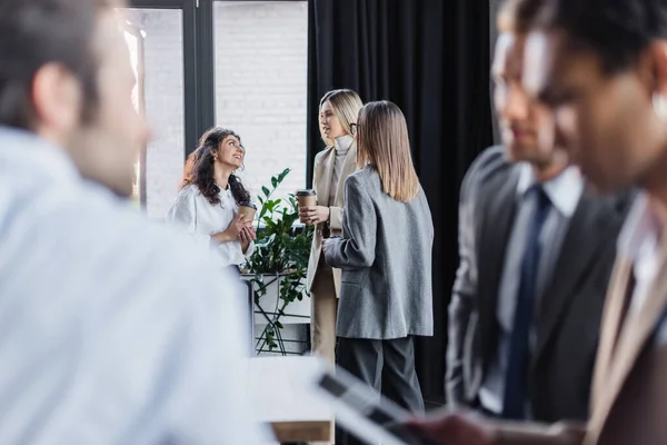 Smiling businesswomen with coffee to go talking near blurred multiethnic businessmen in office — Fotografia de Stock