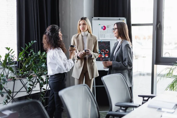 Young businesswomen with coffee to go talking near flip chart with infographics — Foto stock