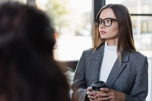 Young businesswoman in eyeglasses holding coffee to go near blurred colleague — Stock Photo