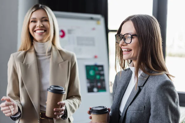Happy businesswomen holding paper cups near blurred flip chart — Stockfoto