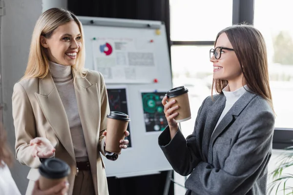Cheerful businesswomen with paper cups talking near blurred flip chart in office — Stockfoto