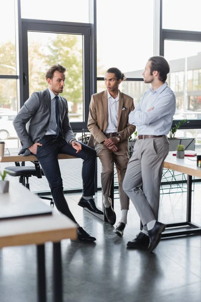 Multicultural businessmen in formal wear sitting on desks while talking in office — Stock Photo