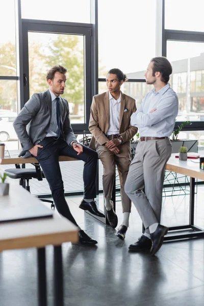 Multicultural businessmen in formal wear sitting on desks while talking in office — Foto stock