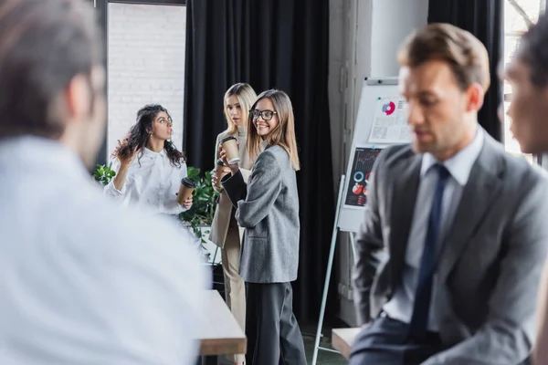 Mujer feliz sosteniendo café para acercarse a colegas y hombres de negocios multiétnicos borrosos - foto de stock