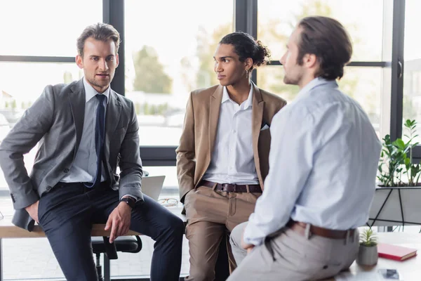 Uomini d'affari multiculturali in abbigliamento formale parlando mentre seduti sulle scrivanie in ufficio — Foto stock