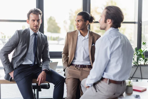 Multicultural businessmen in formal wear talking while sitting on desks in office — Stock Photo