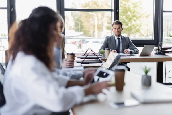 Blurred businesswomen sitting at desk near young businessman in formal wear — Foto stock