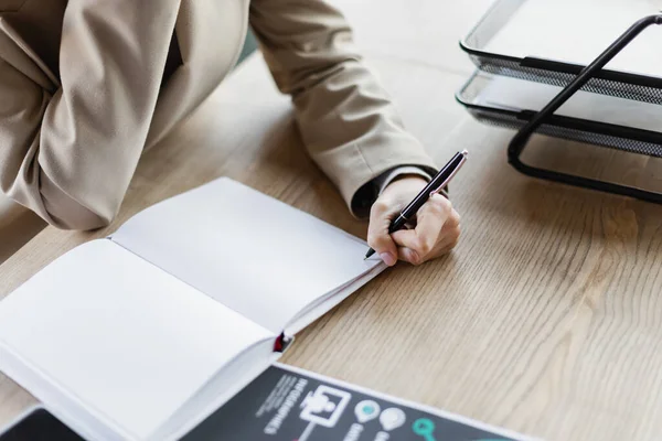 Cropped view of businesswoman holding pen near blank notebook in office — Fotografia de Stock