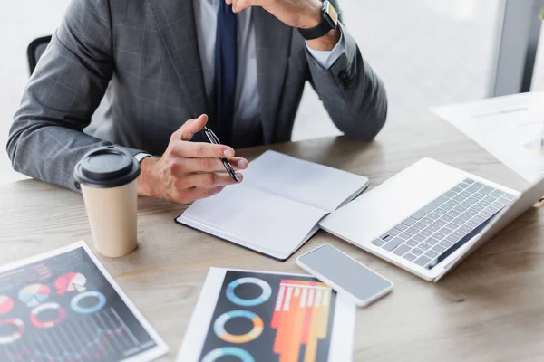 Vista parcial del hombre de negocios que sostiene la pluma cerca del cuaderno vacío, computadora portátil y gráficos en el escritorio - foto de stock