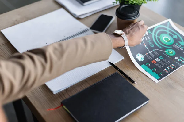 Cropped view of african american businessman taking infographics near notebooks, coffee to go and smartphone on desk — Stock Photo