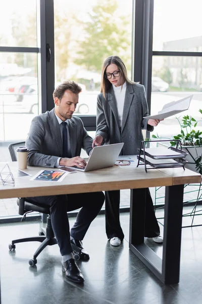 Бізнес-леді в окулярах вказує на ноутбук біля колеги, що працює в офісі — стокове фото