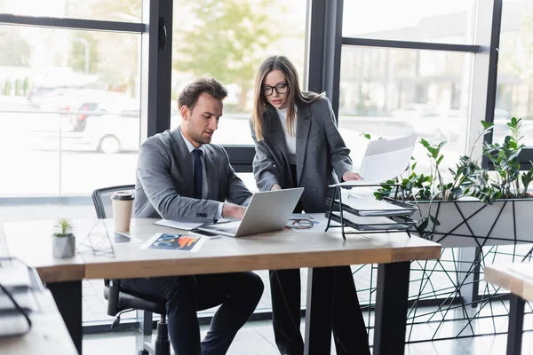 Young businesswoman holding folder while standing near colleague working on laptop — Stock Photo