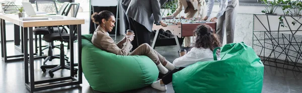 Interracial business people sitting in bag chairs near colleagues playing table football, banner — Stockfoto