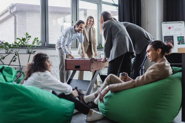 Fröhliche Geschäftsleute, die neben verschwommenen interrassischen Kollegen Tischfußball spielen, die in Tütenstühlen sitzen — Stockfoto