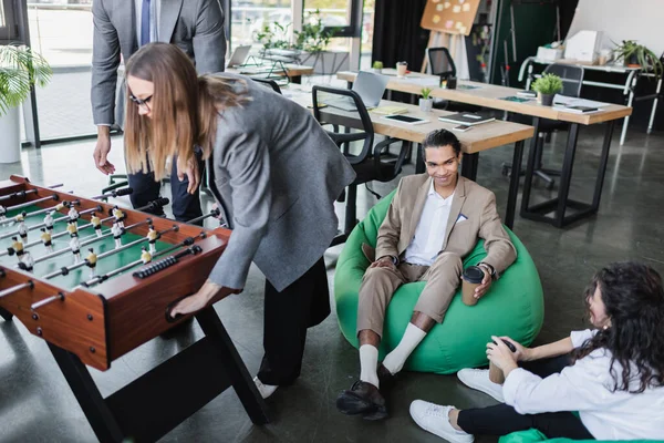 Gente de negocios interracial sentada en sillas de bolsa cerca de colegas jugando futbolín en la oficina - foto de stock