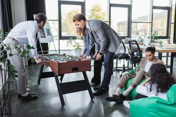 Junge Geschäftsleute spielen Tischfußball in der Nähe multiethnischer Kollegen, die in Tütenstühlen sitzen — Stockfoto