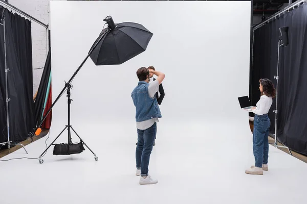 Director de arte portatil con pantalla en blanco cerca de fotógrafo y modelo trabajando en estudio fotográfico - foto de stock