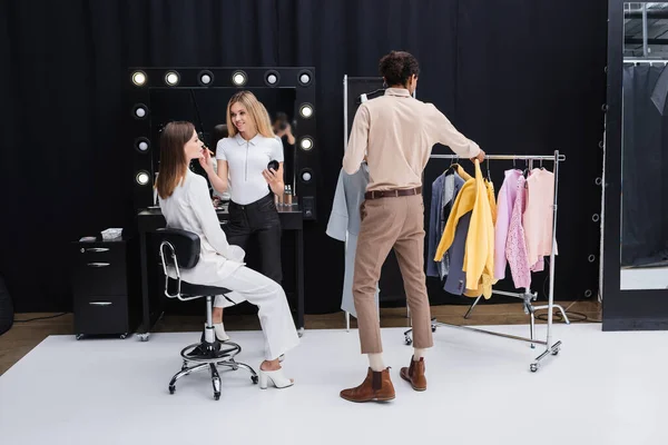 Afroamericano estilista elegir ropa mientras maquillaje artista haciendo visage a modelo en vestidor - foto de stock