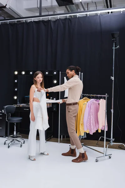 Vue pleine longueur du styliste afro-américain habiller jeune femme brune dans le studio photo — Photo de stock