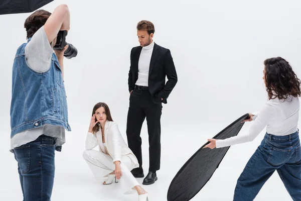 Trendy models posing near photographer and assistant with reflector on white — Stock Photo