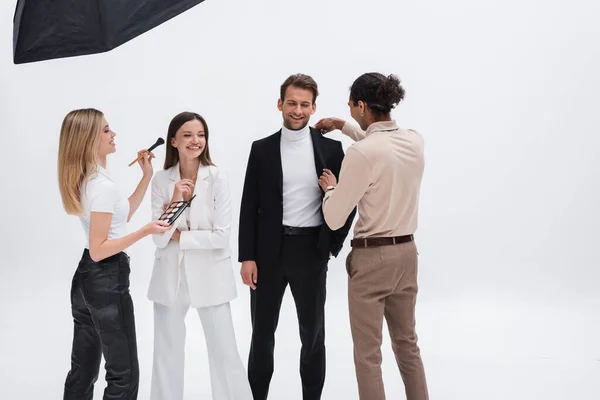 Stylish models smiling near makeup artist and african american stylist preparing them to photo session on white — Stock Photo