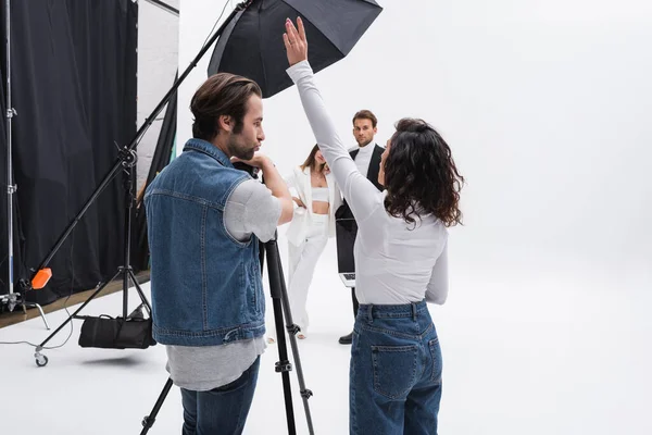 Director de arte de pie con la mano levantada mientras habla con el fotógrafo cerca de modelos en el estudio de fotos - foto de stock