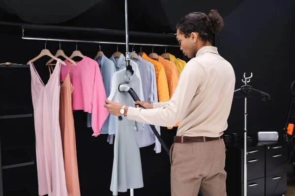 Vestido de planchar afroamericano con vaporizador cerca de rack con ropa en vestidor - foto de stock