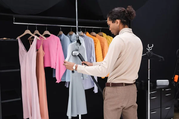 Robe de repassage costumier afro-américain avec vapeur près du rack avec des vêtements dans le vestiaire — Photo de stock