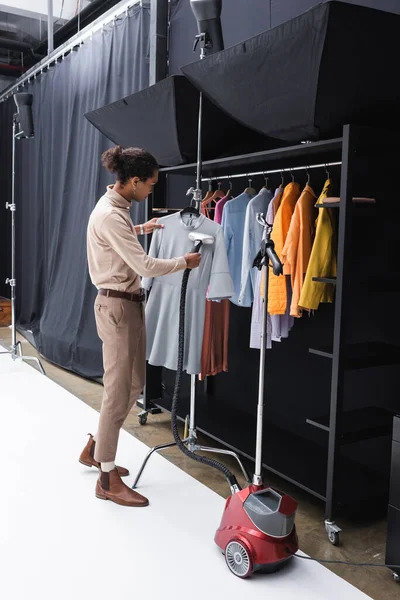 Young african american costumier ironing clothes with steamer in dressing room — Stock Photo