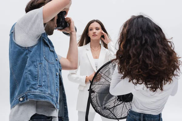 Brunette femme en costume blanc posant près photographe et assistant avec ventilateur électrique isolé sur blanc — Photo de stock